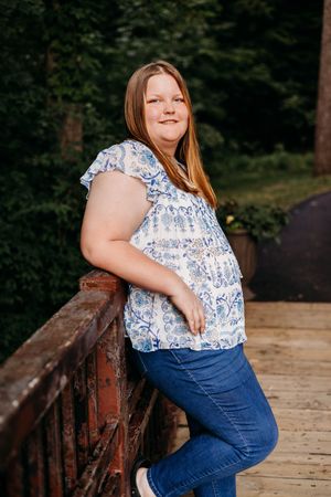 A person in a blue floral top and jeans leans casually against a wooden railing in an outdoor setting.