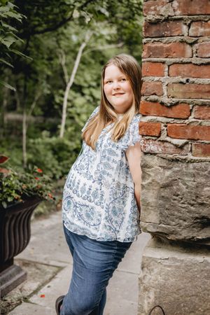 A person in a blue and white patterned top and jeans leans against a rustic brick wall surrounded by greenery.