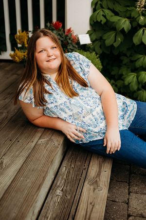 A person in a blue floral blouse and jeans relaxes on wooden deck steps with greenery in the background.
