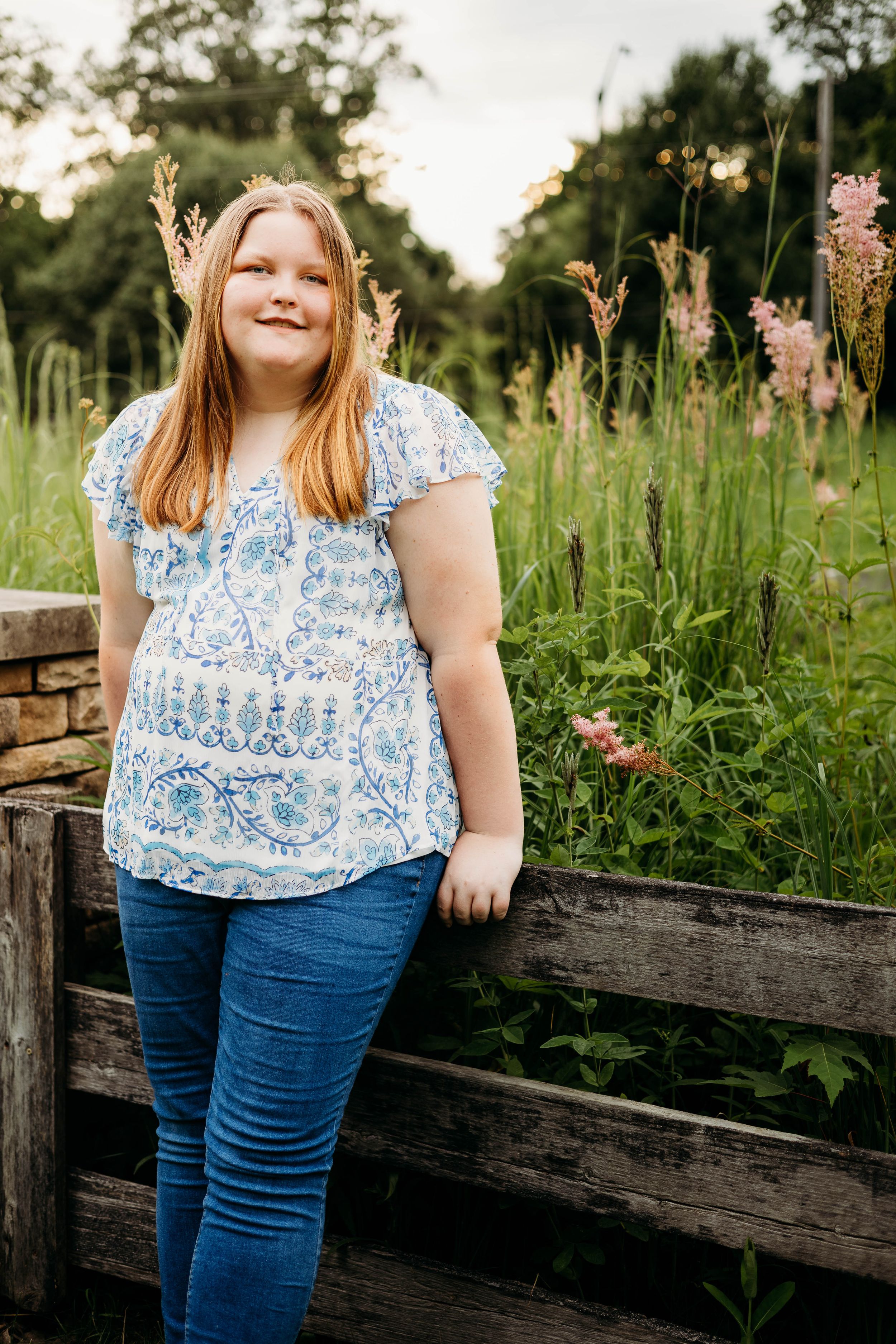 Standing by a rustic wooden fence at sunset in a floral print top and jeans among wildflowers in a meadow.