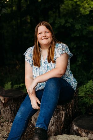 Someone in a floral top and blue jeans sits casually on a tree stump in a wooded outdoor setting.