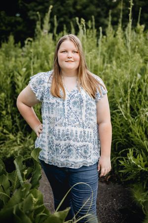 Person in blue jeans and patterned short-sleeve top poses outdoors against lush green foliage on a sunny day.