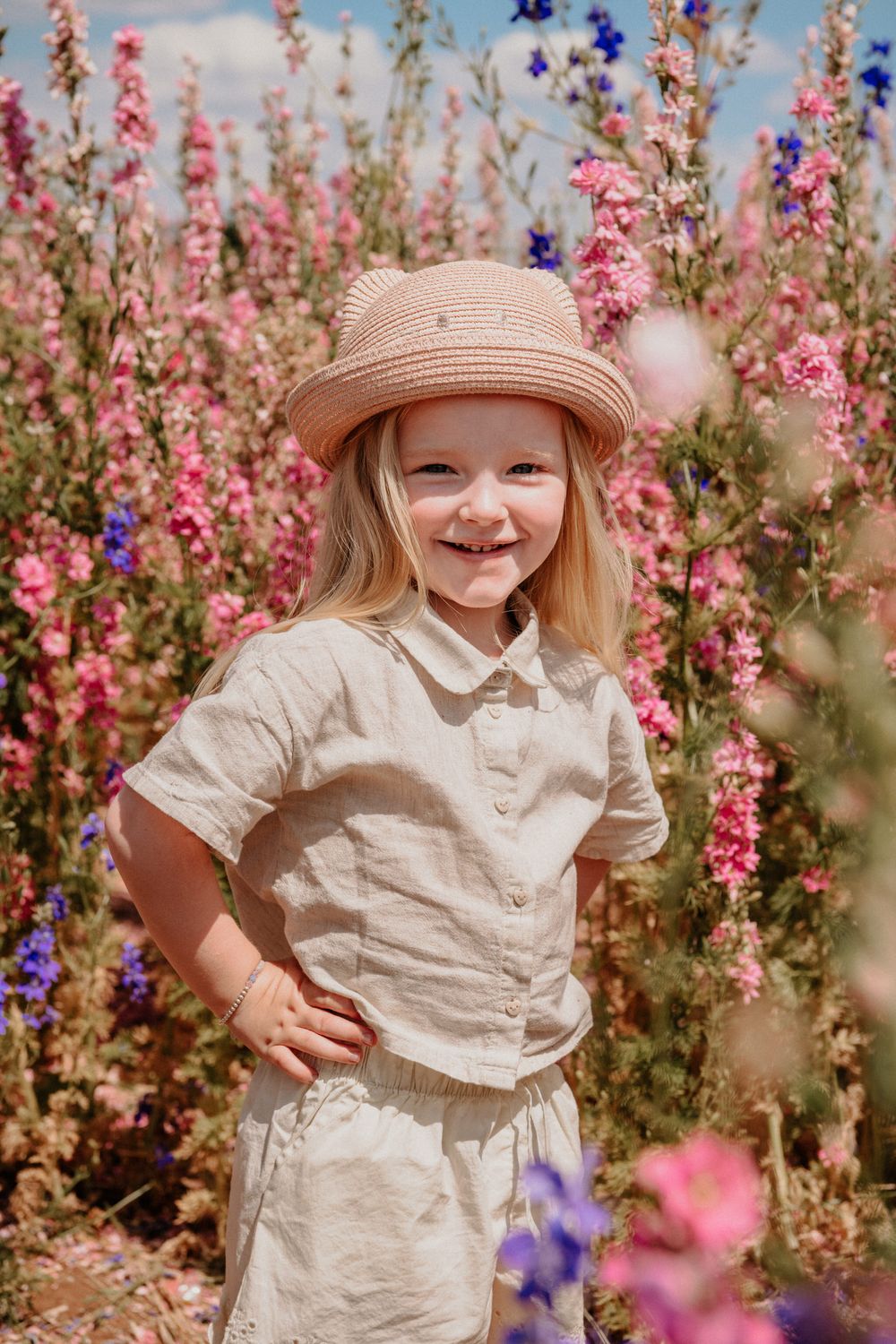 A child wearing a straw hat and white outfit plays joyfully among pink and purple wildflowers in a garden.