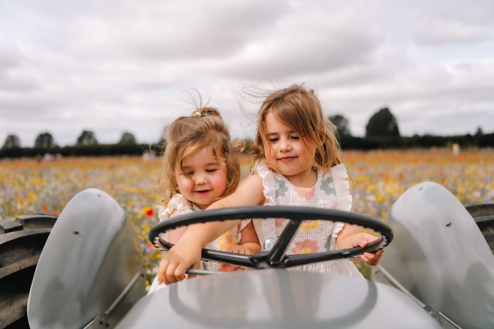 Two young girls smiling while sitting in a vintage toy car in a colorful flower field.
