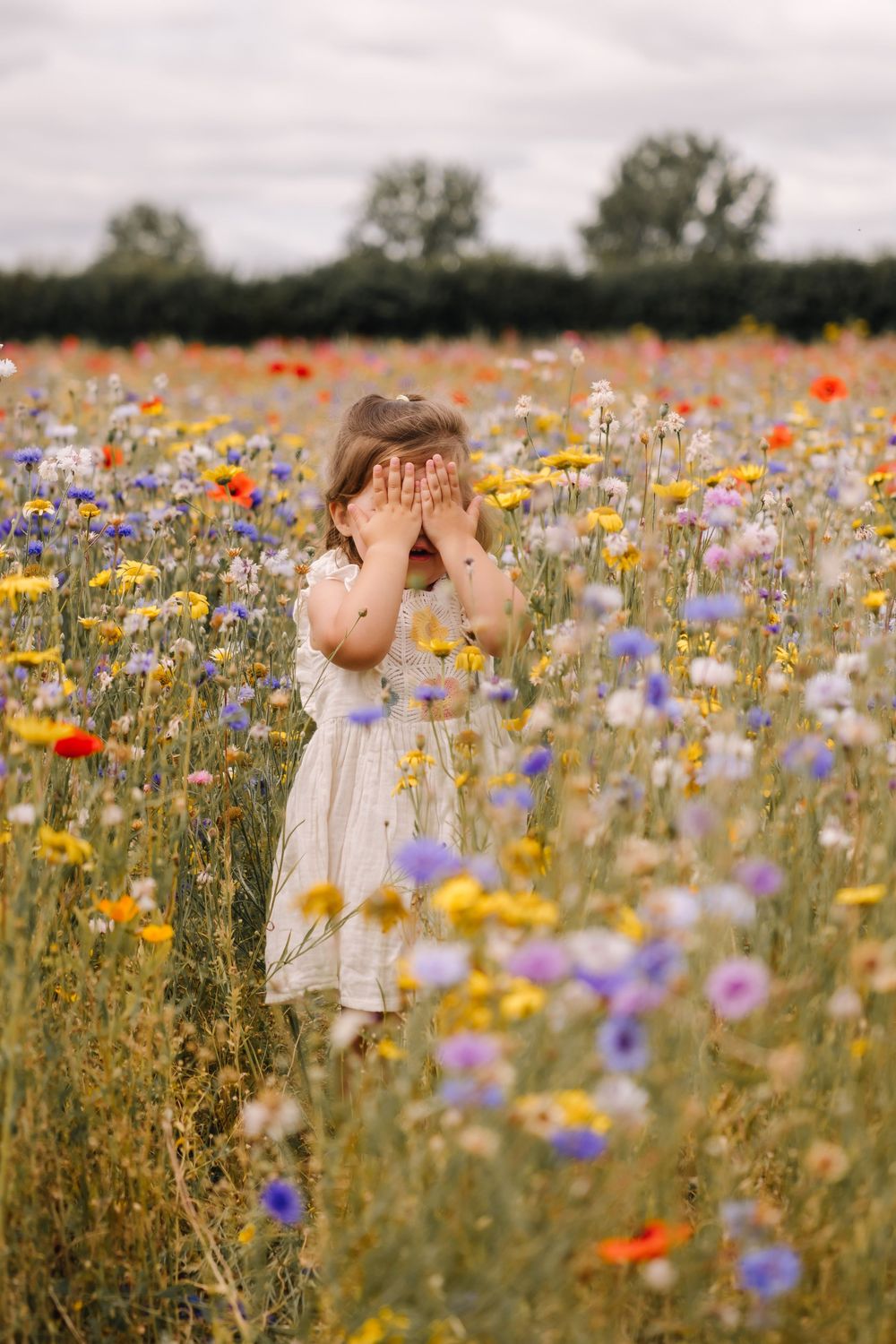 Young girl in white dress covering her face with hands standing in a vibrant wildflower field with poppies.