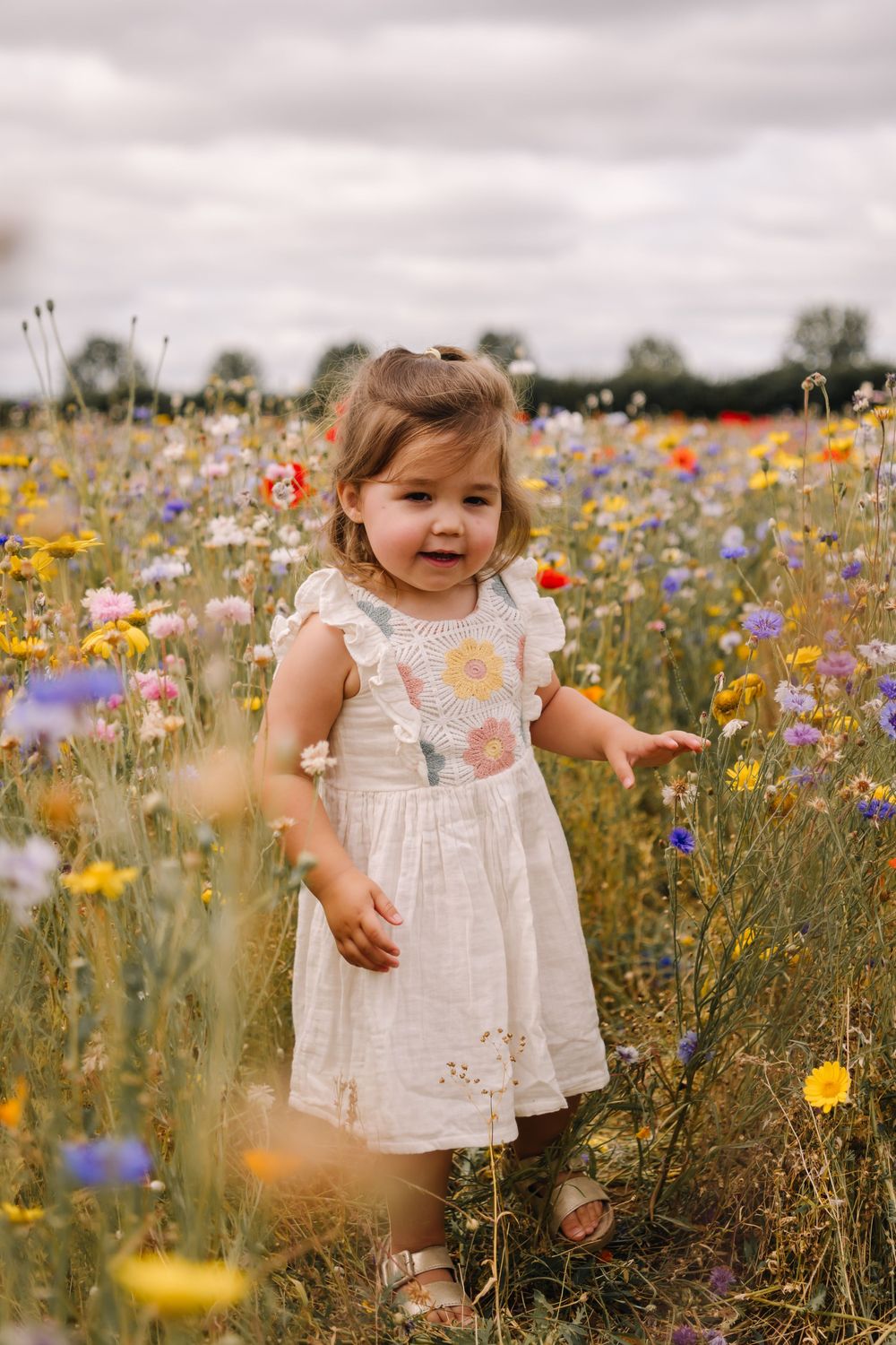 Toddler girl in white floral dress standing in a vibrant wildflower meadow with colorful blooms on a cloudy day.