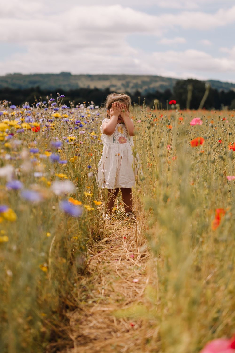 Young girl in white dress covering her face standing in a vibrant wildflower field with poppies and cornflowers.