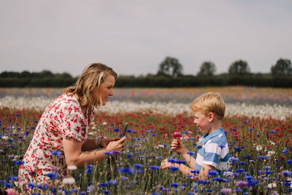 A woman in a floral dress bends down to a young boy holding flowers in a vibrant wildflower field.
