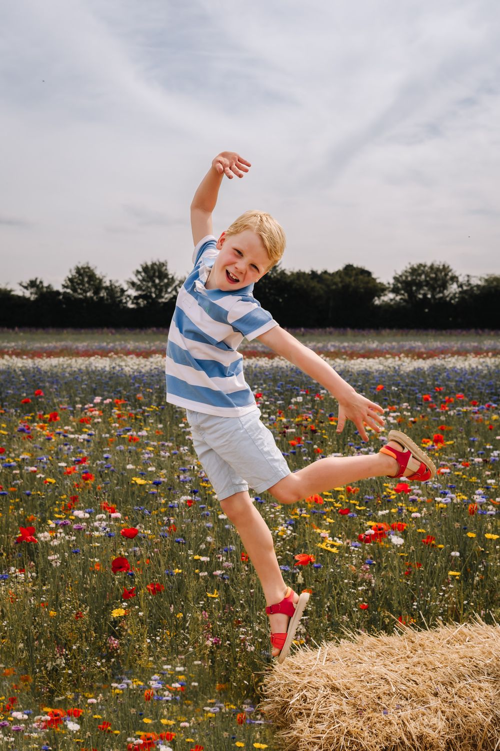 A joyful blonde boy jumps in a colorful wildflower field, wearing a striped shirt and red sandals on a cloudy day.