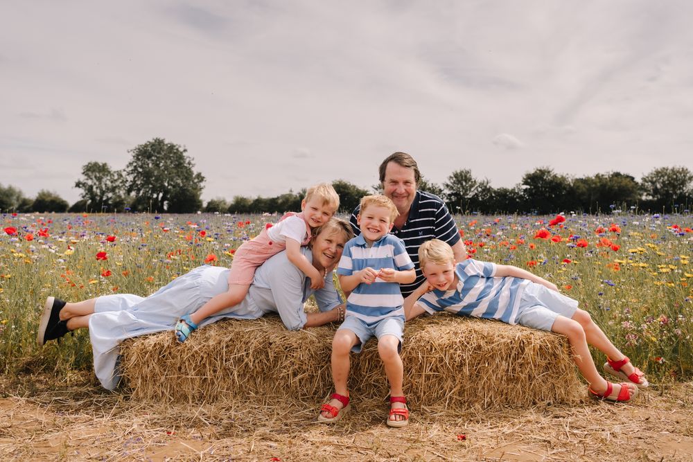 Family of five posing on a hay bale in a colorful wildflower field with red poppies on a cloudy day.