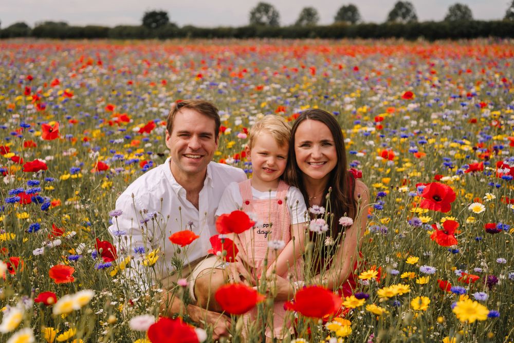 Family of three smiling in a vibrant wildflower field with red poppies and colorful blooms on a sunny day.