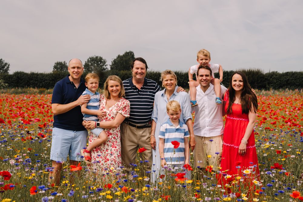 Family group portrait in a vibrant wildflower meadow with red poppies, adults and children smiling outdoors.
