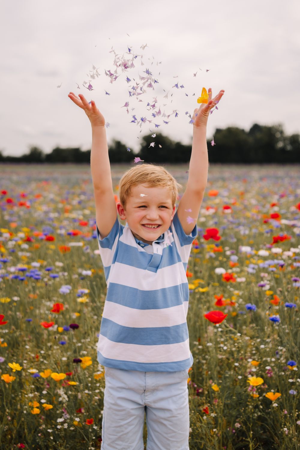 A joyful blond boy tosses colorful flower petals in the air while standing in a vibrant wildflower meadow.