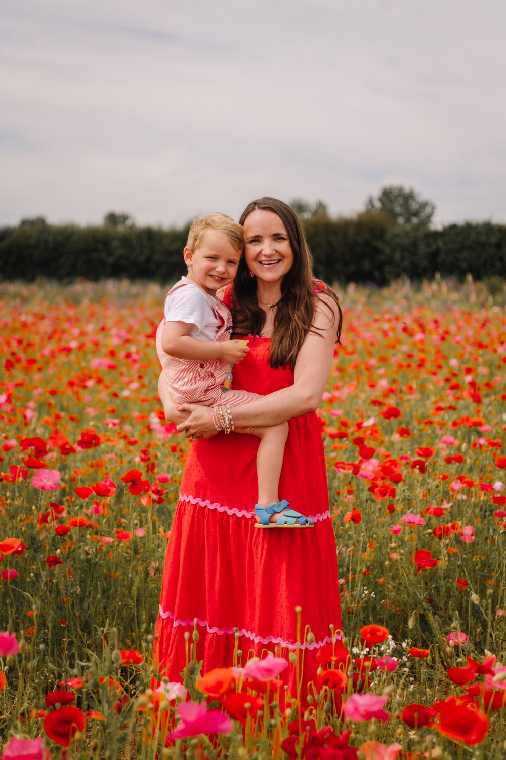 Woman in red dress holding a young child in a colorful poppy field on an overcast day.