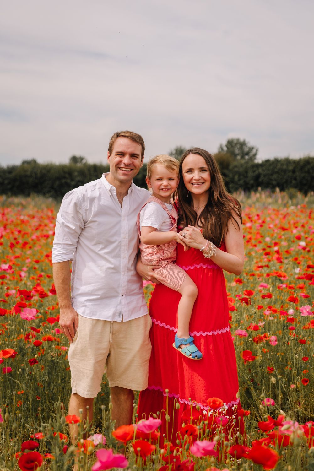 Happy family of three posing in a vibrant red poppy field, parents smiling with young child in matching red outfits.