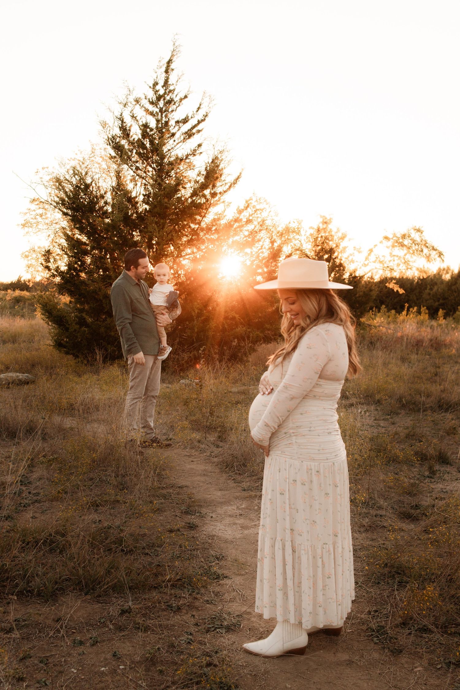 A couple poses in a field at sunset with evergreen trees in the background.