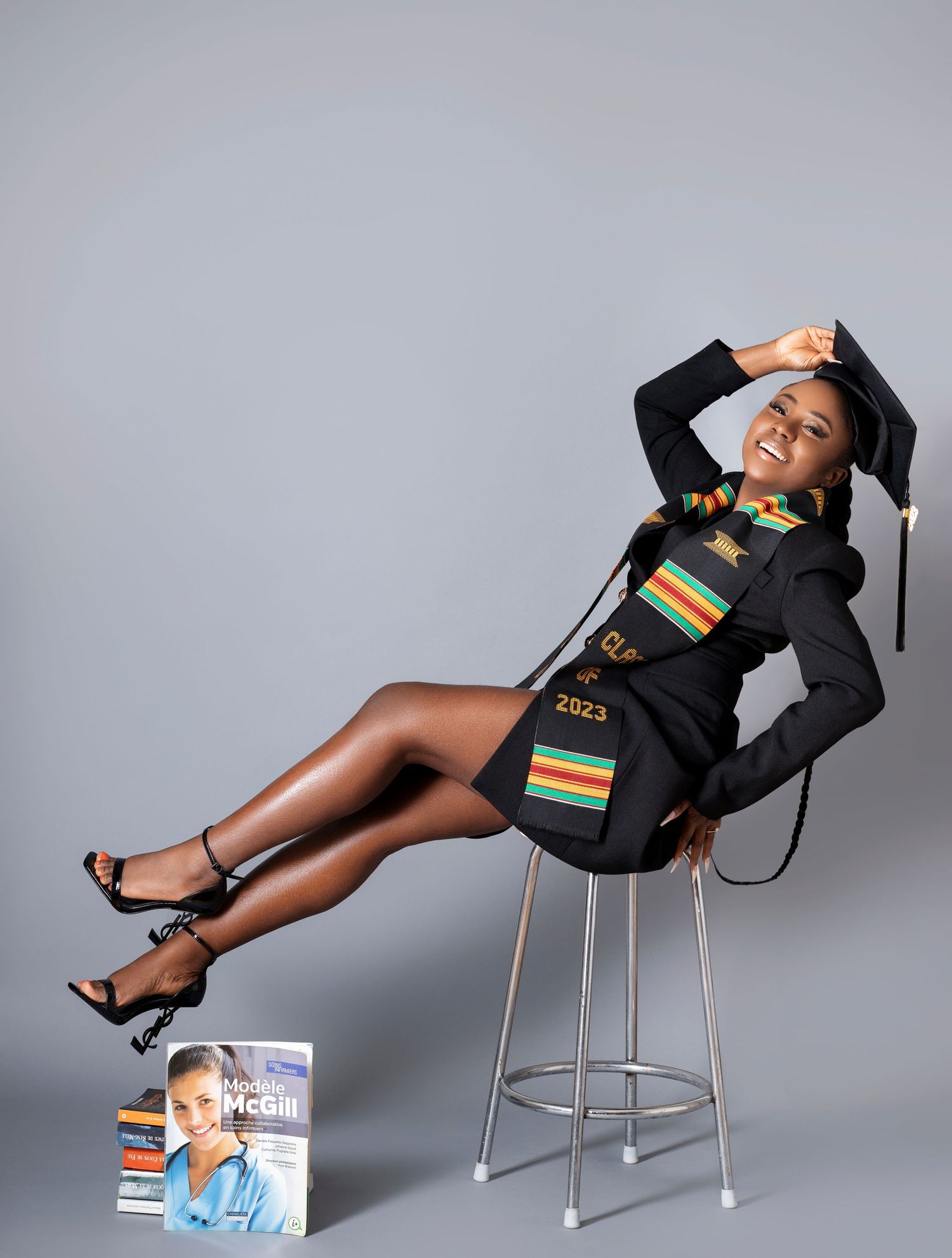 Graduate in academic regalia poses dramatically on stool in studio setting.