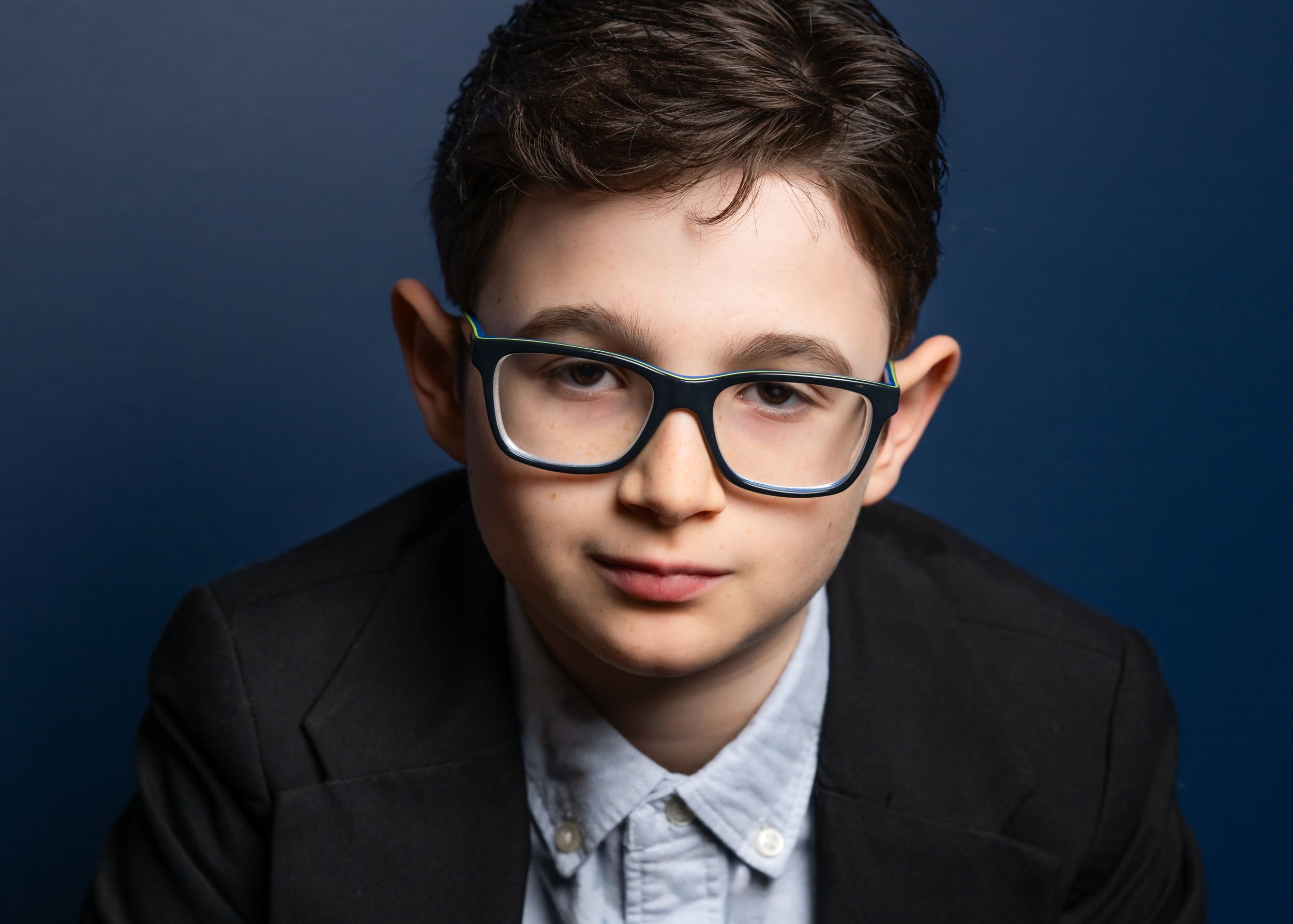 A professional headshot against deep blue background featuring black-rimmed glasses.