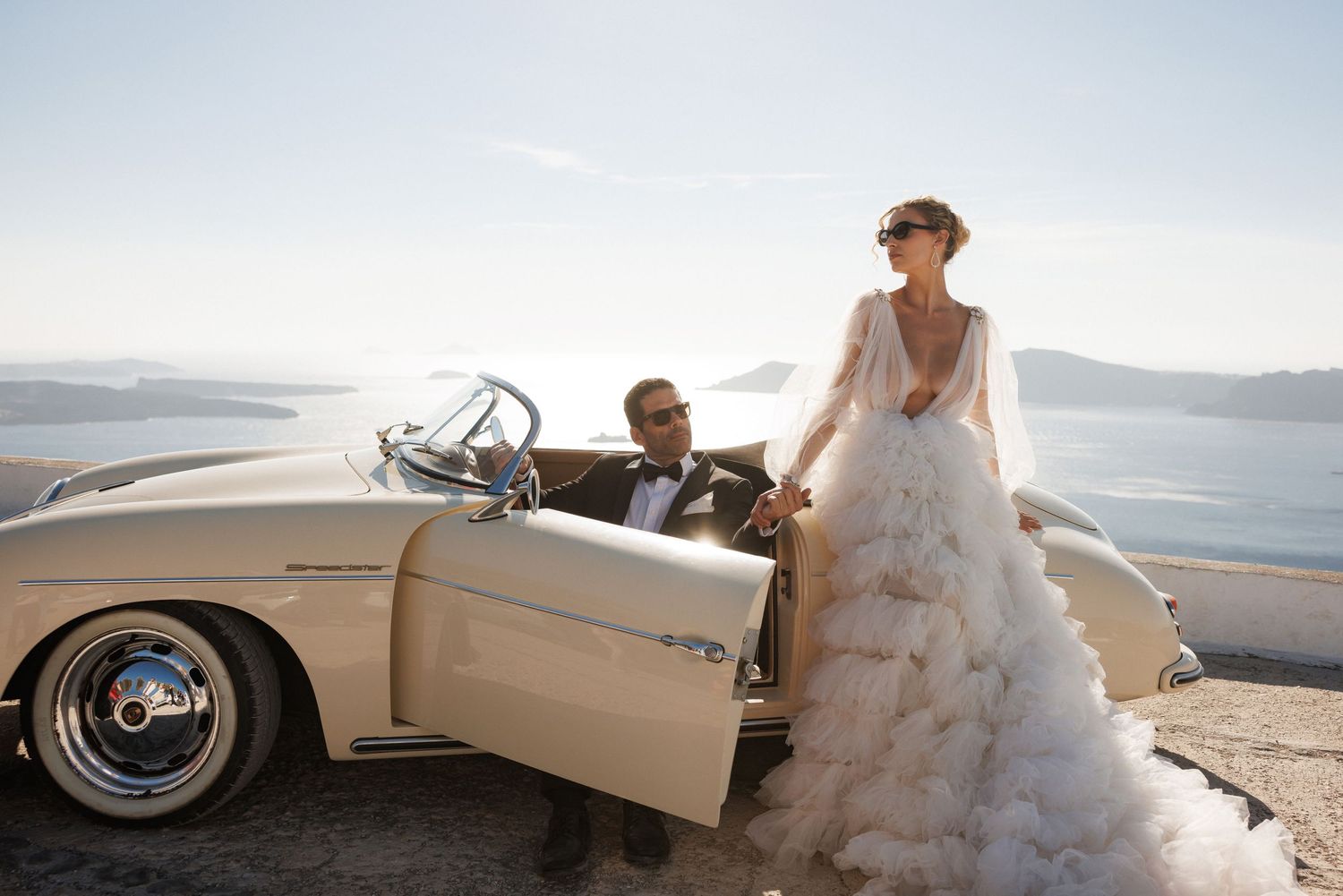 A couple in wedding attire pose with a classic cream-colored convertible car along a scenic coastal road.