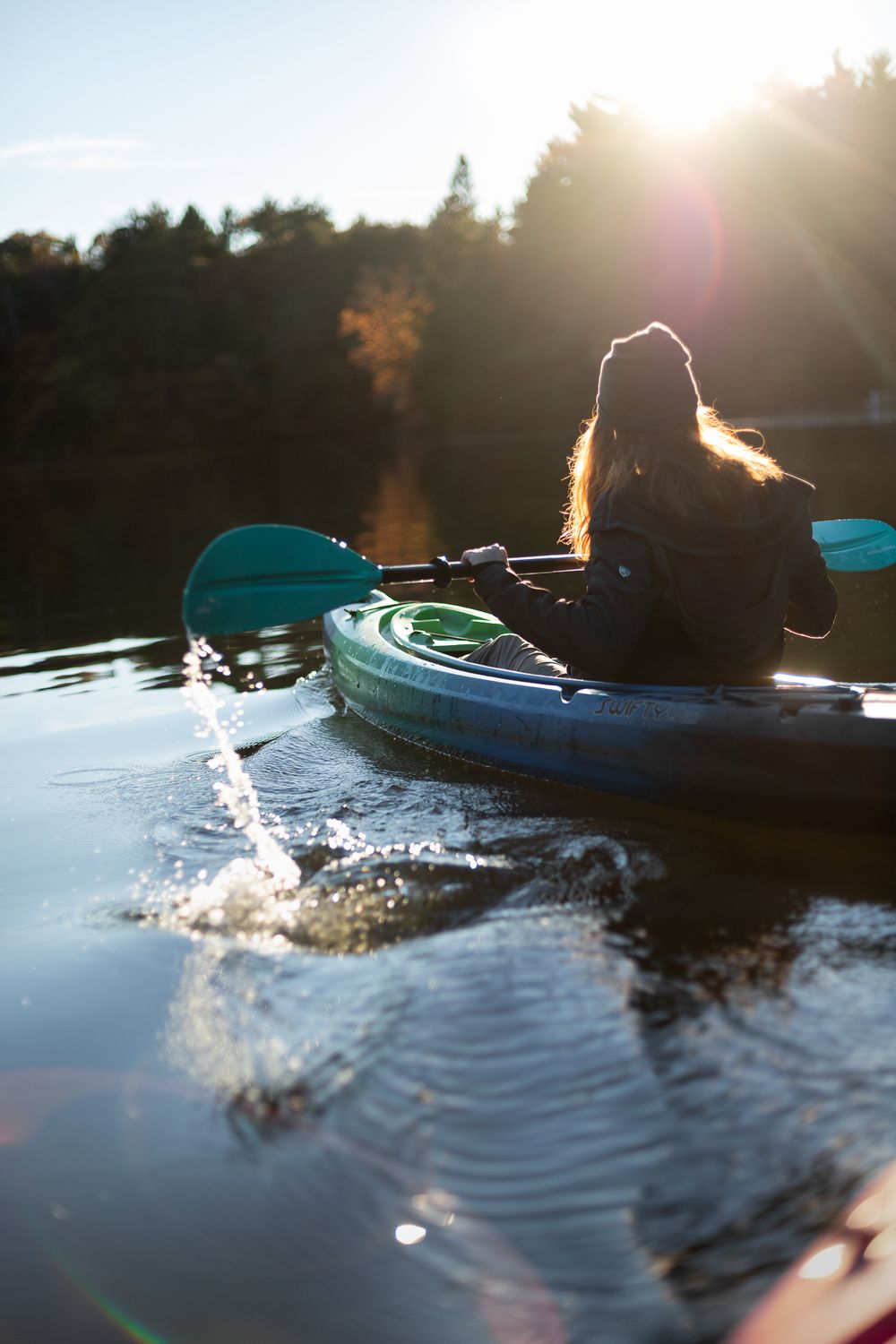 girl in beanie hat kayaking through water at sunset making splashes with paddle