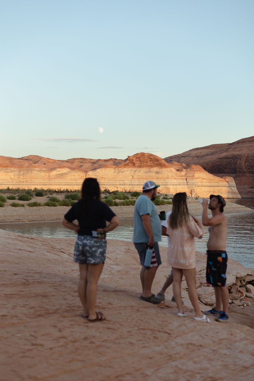 group of people standing on pink sandy rocks overlooking water and canyons in distance