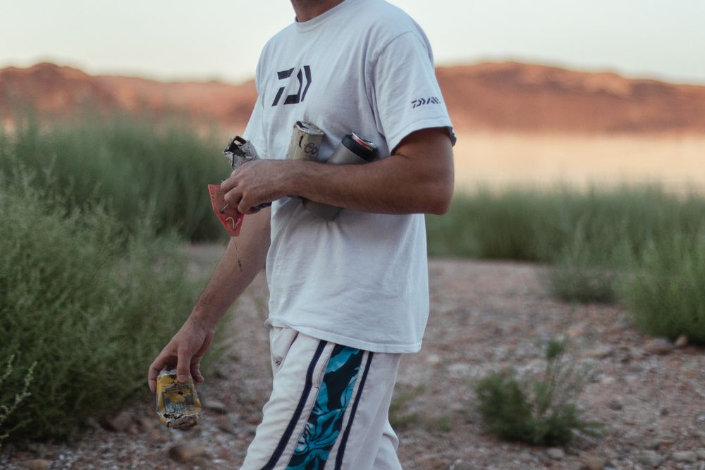 man in white shirt carrying an arm full of garbage out of sandy rocky area in desert