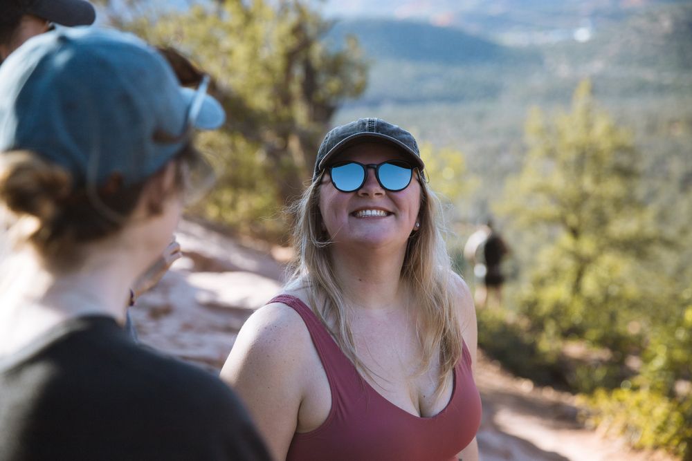 lady smiling at camera wearing ball cap and sunglasses with trees and rocks in background