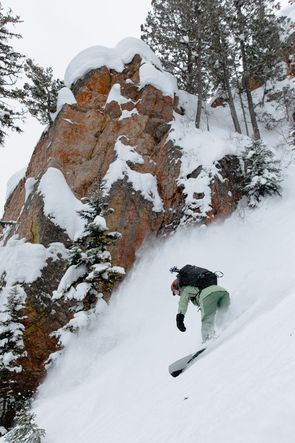 person in green snowboarding downhill alongside snow covered cliff and trees