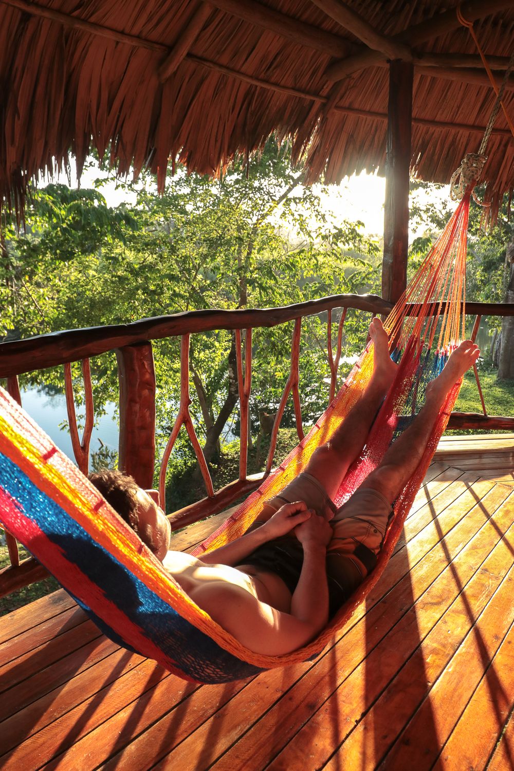 man lounging in colorful hammock on porch of bungalow overlooking sunset and a river in the jungle below