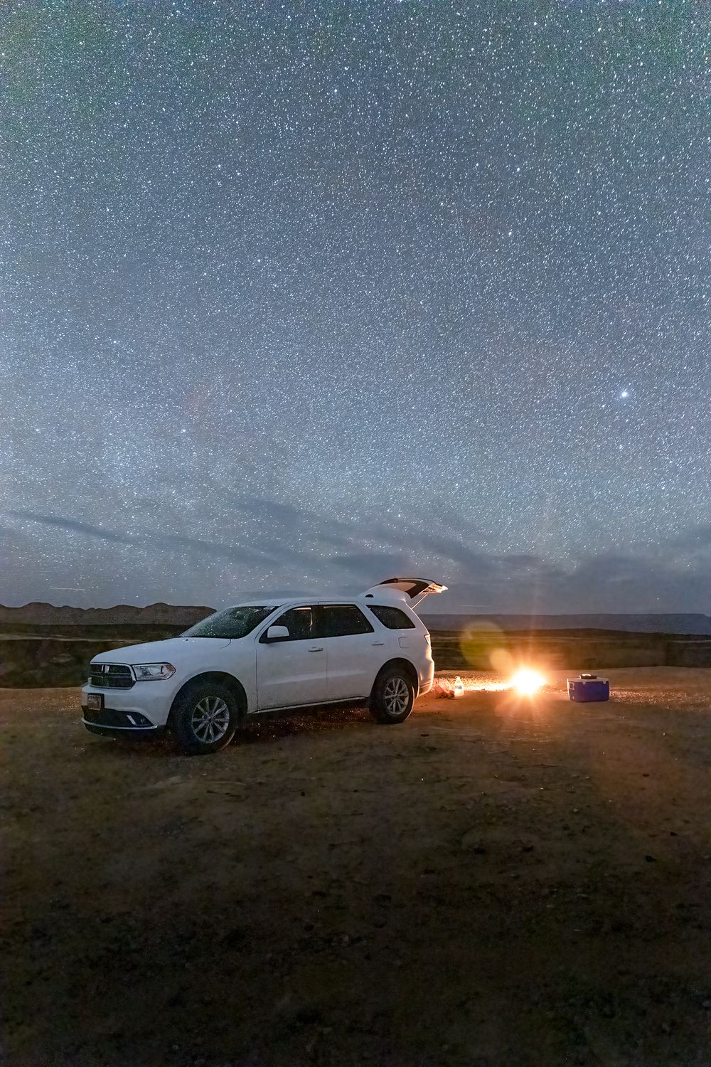 white SUV parked on a ledge at night with stars above and tailgate open with a fire and cooler beside it