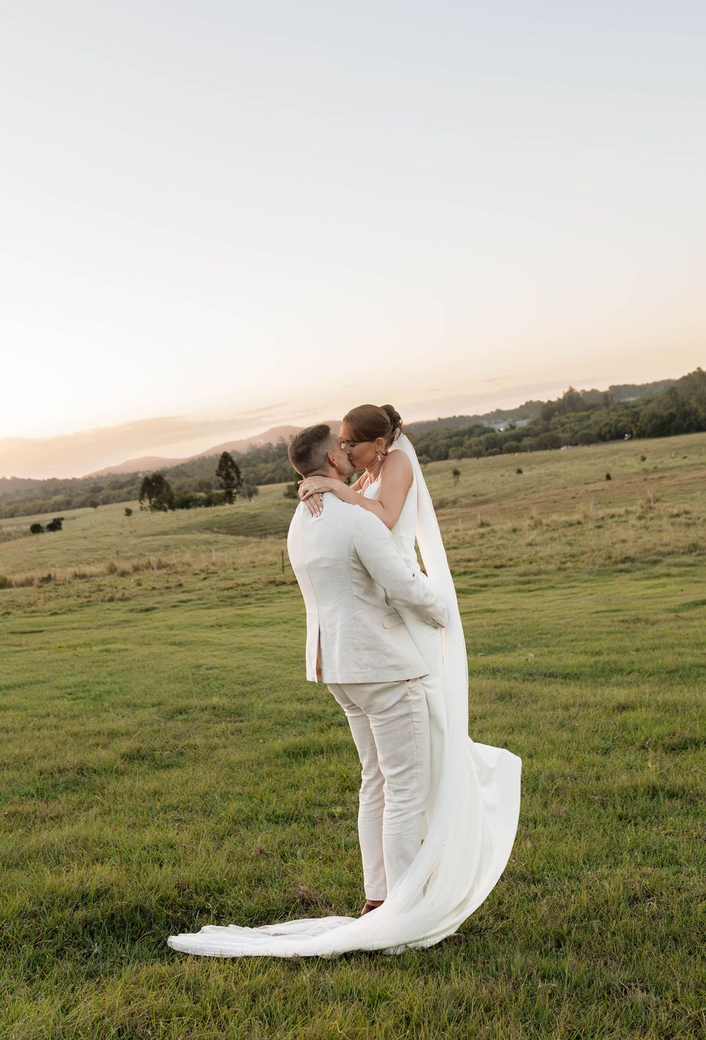 A bride and groom share a romantic kiss in a lush green field at sunset, dressed in elegant white wedding attire.