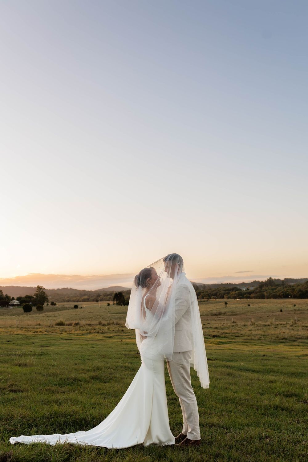 Bride and groom share a kiss under veil at sunset in a green field with rolling hills backdrop.