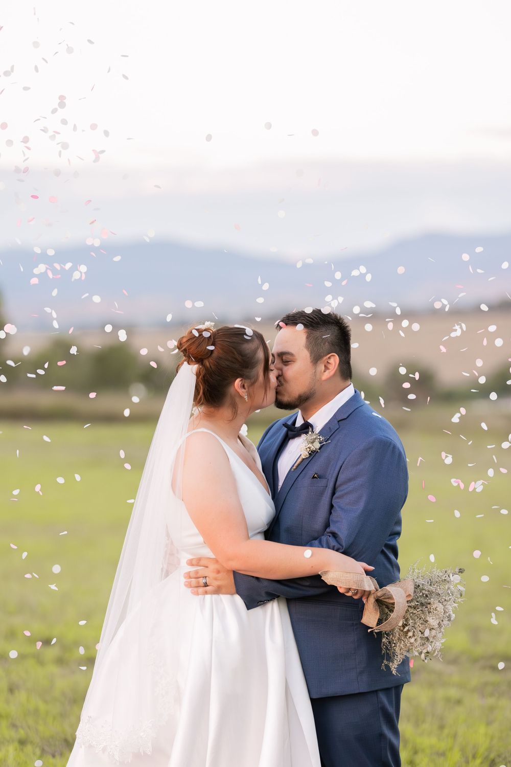 Romantic wedding kiss with confetti falling in sunlit field with mountain backdrop.