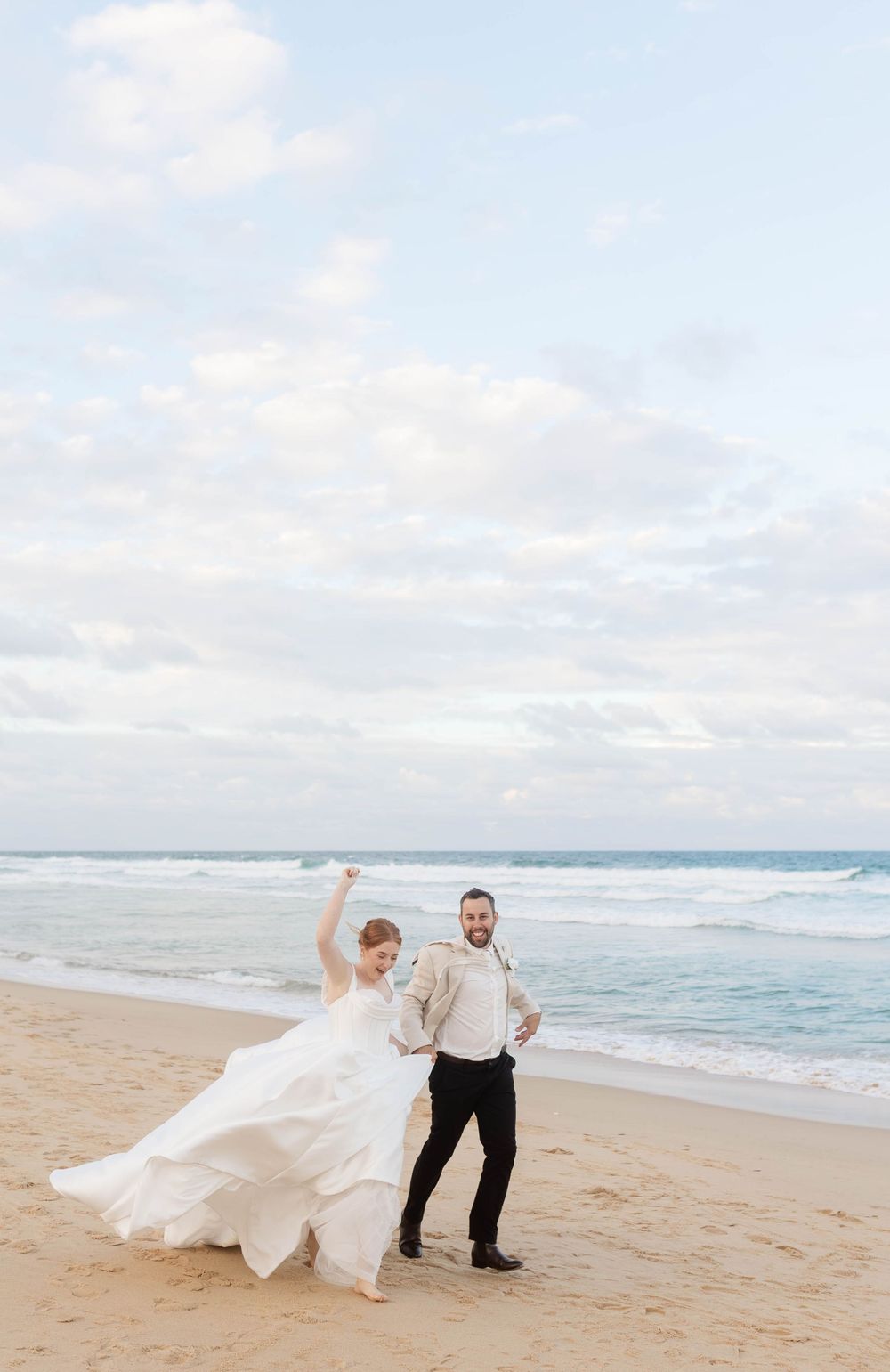 Joyful bride and groom celebrating on a sandy beach with ocean waves behind them.