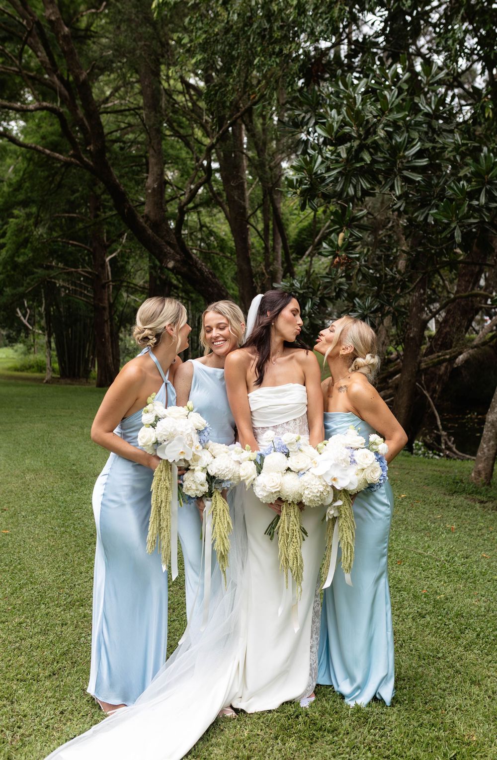 Bride in white gown with three bridesmaids in light blue satin dresses holding white floral bouquets outdoors.