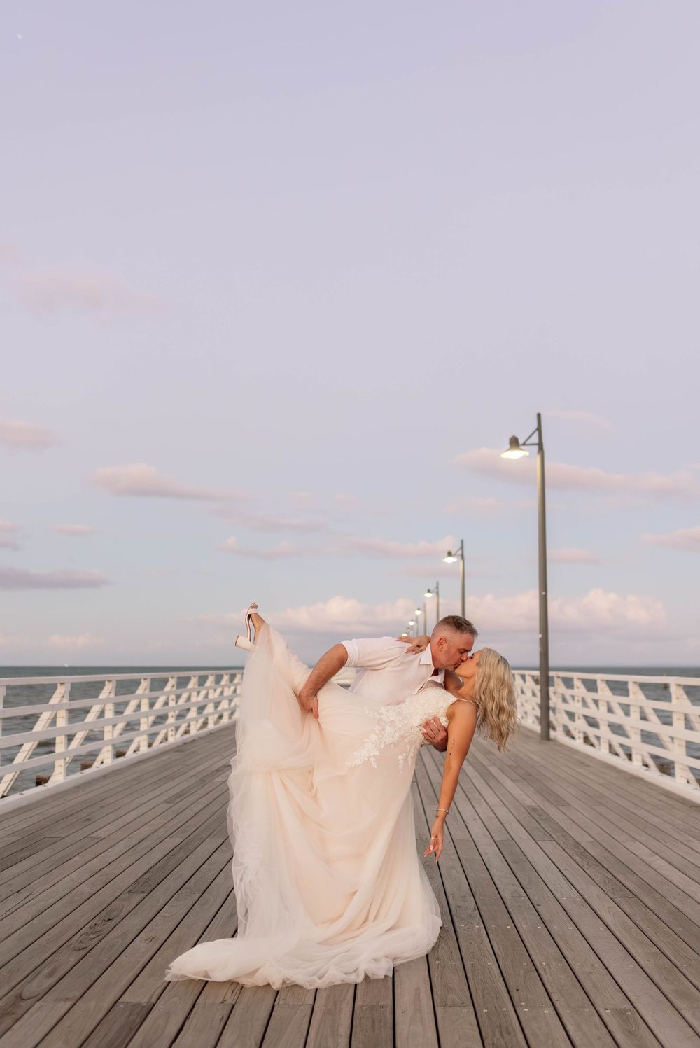 Bride and groom share a romantic dip kiss on a wooden pier at dusk, with ocean views and lamp posts.