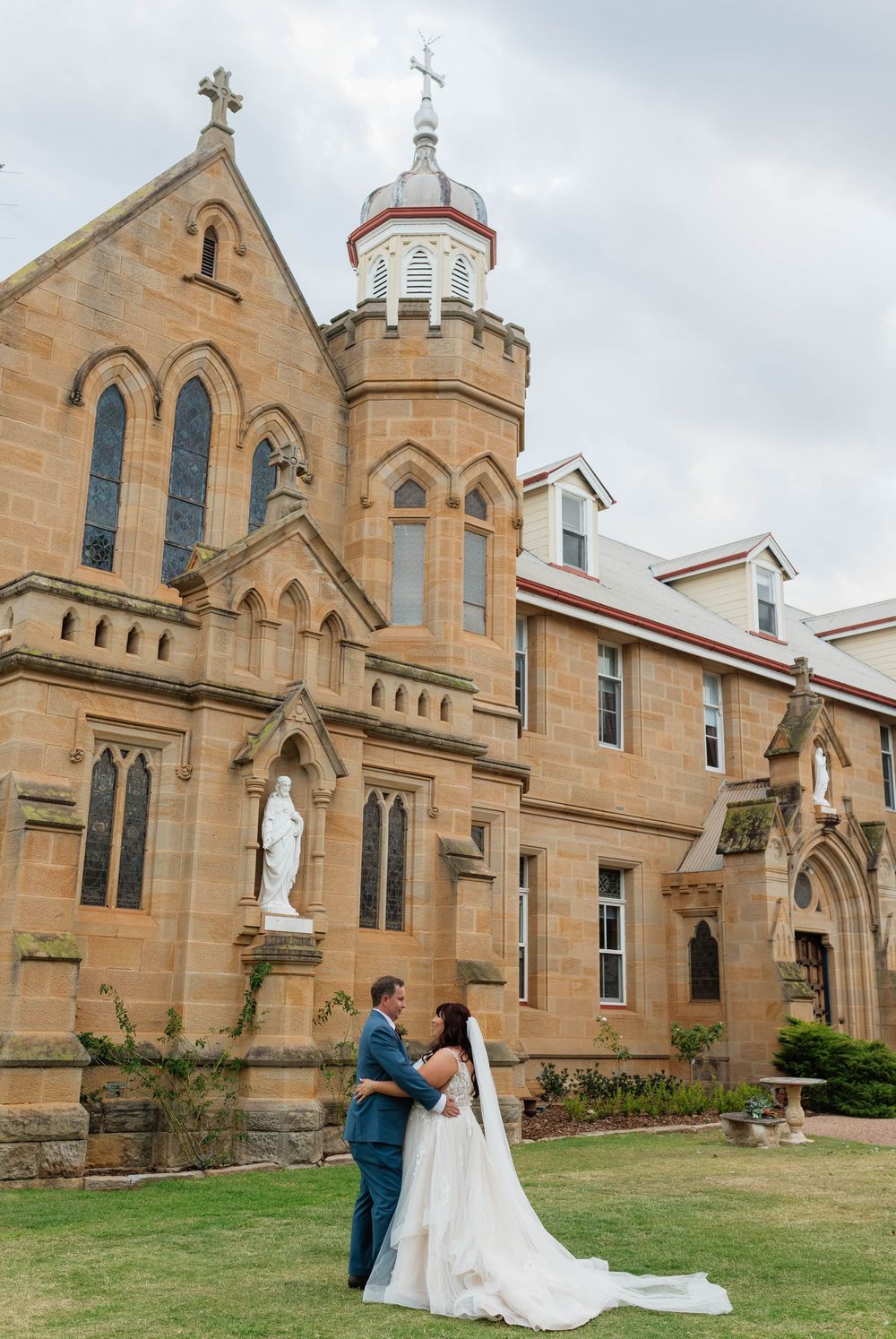 Bride and groom embrace outside a historic sandstone church with Gothic architecture and religious statues on their wedding day.