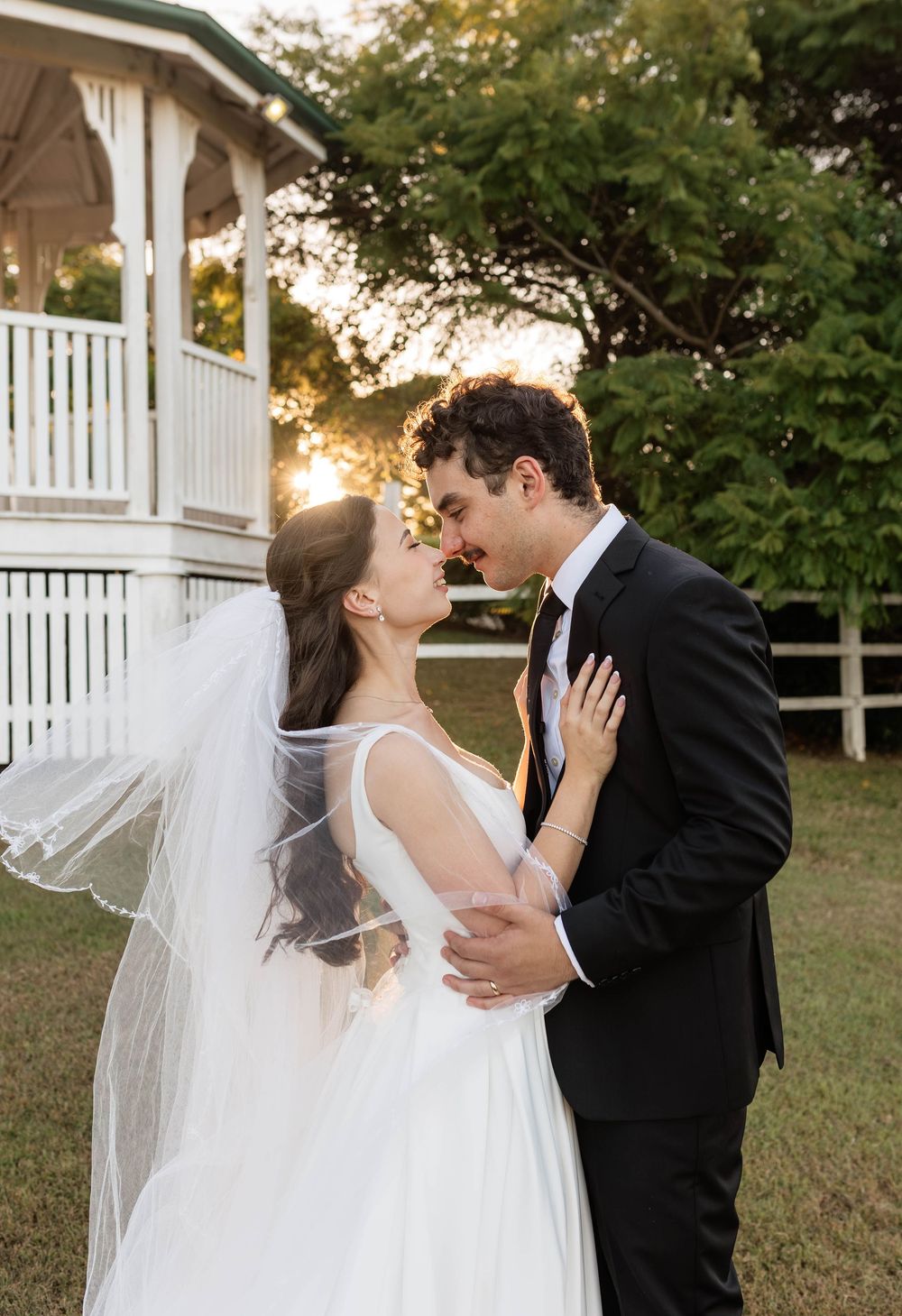 A bride and groom share a romantic moment at golden hour near a white gazebo, with the veil flowing softly in the breeze.