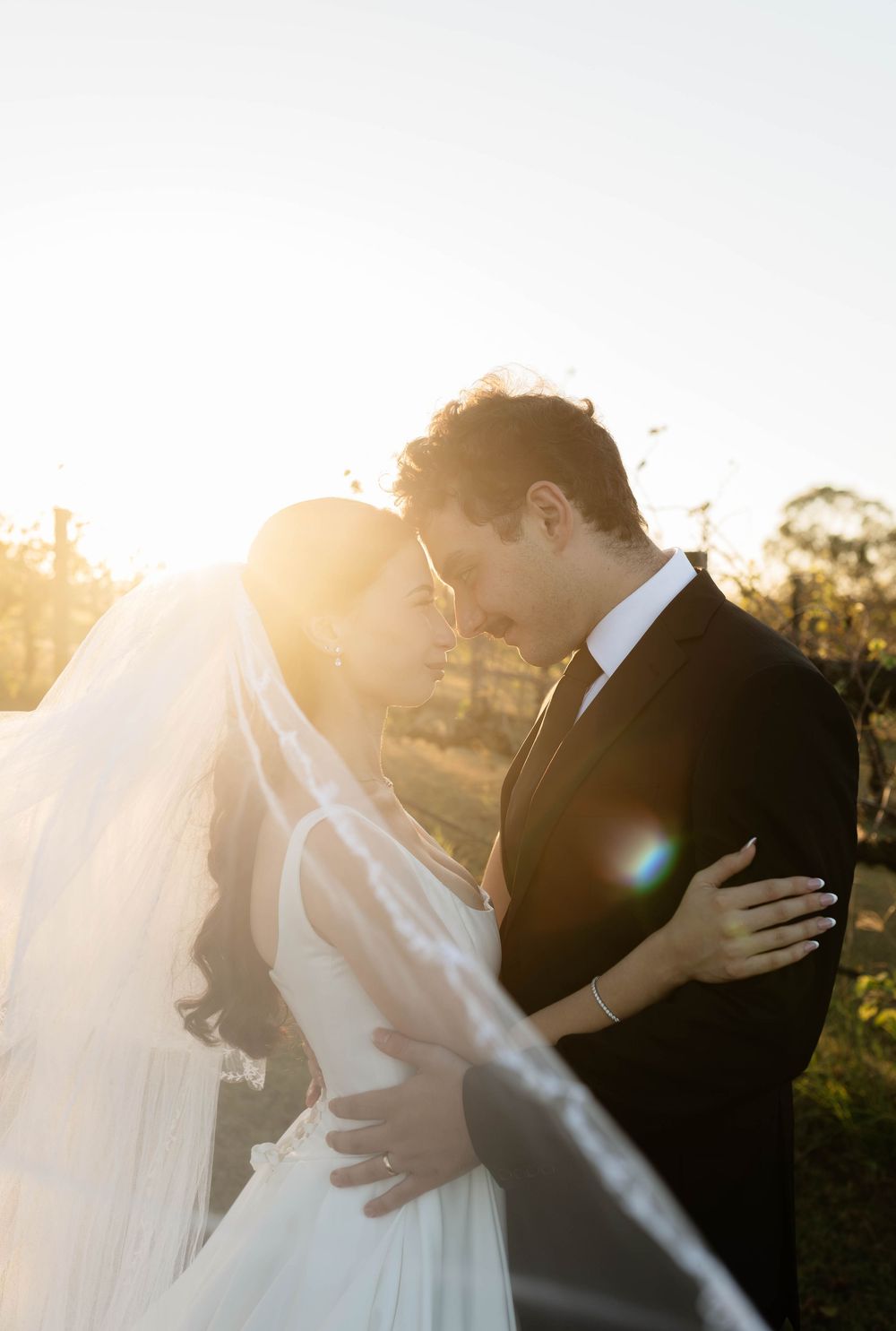 Bride and groom share an intimate moment at golden hour, with wedding veil flowing and warm sunlight glowing behind them.