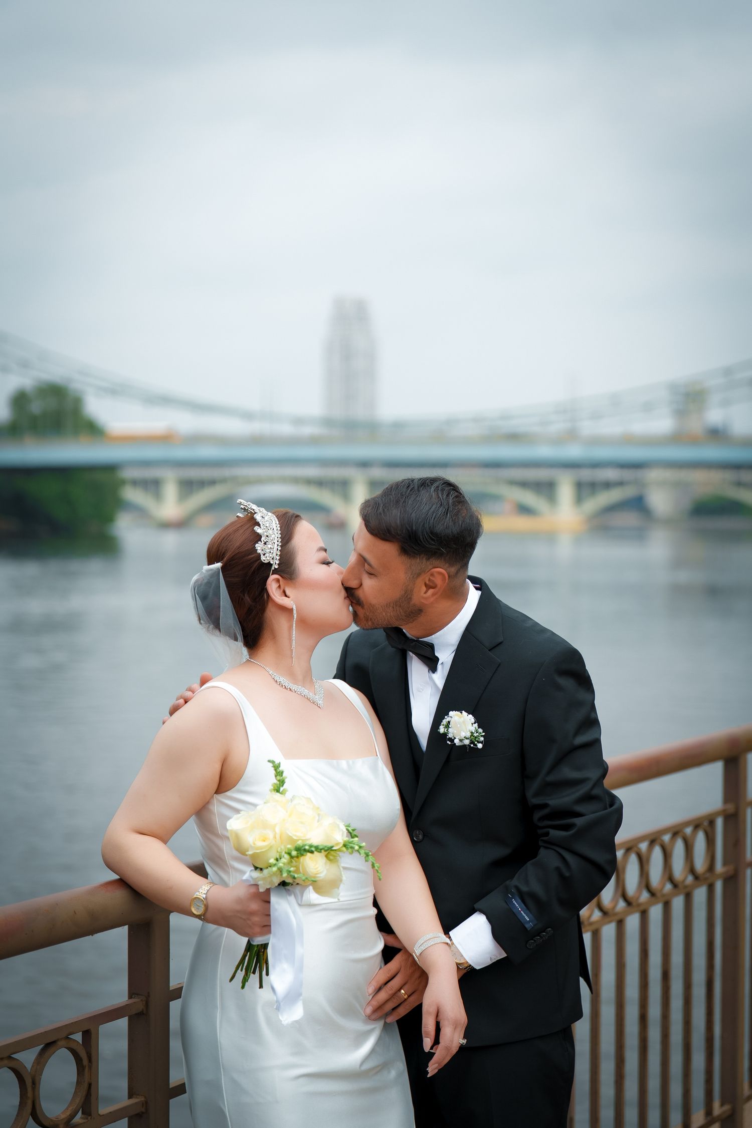 A newlywed couple shares romantic moments along a waterfront with a bridge visible in the background.
