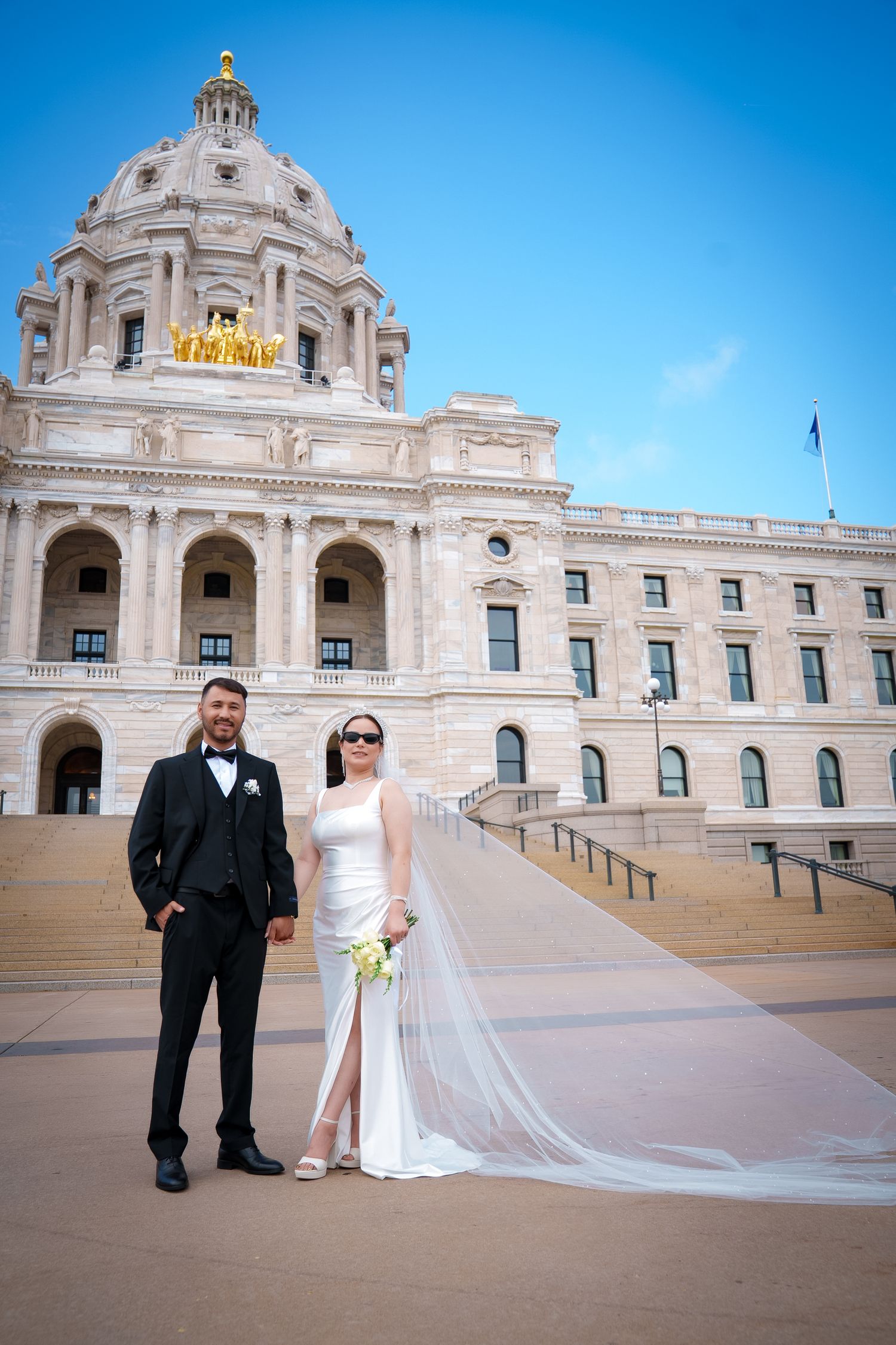 Wedding couple poses on the steps of the Minnesota State Capitol building under bright blue skies.
