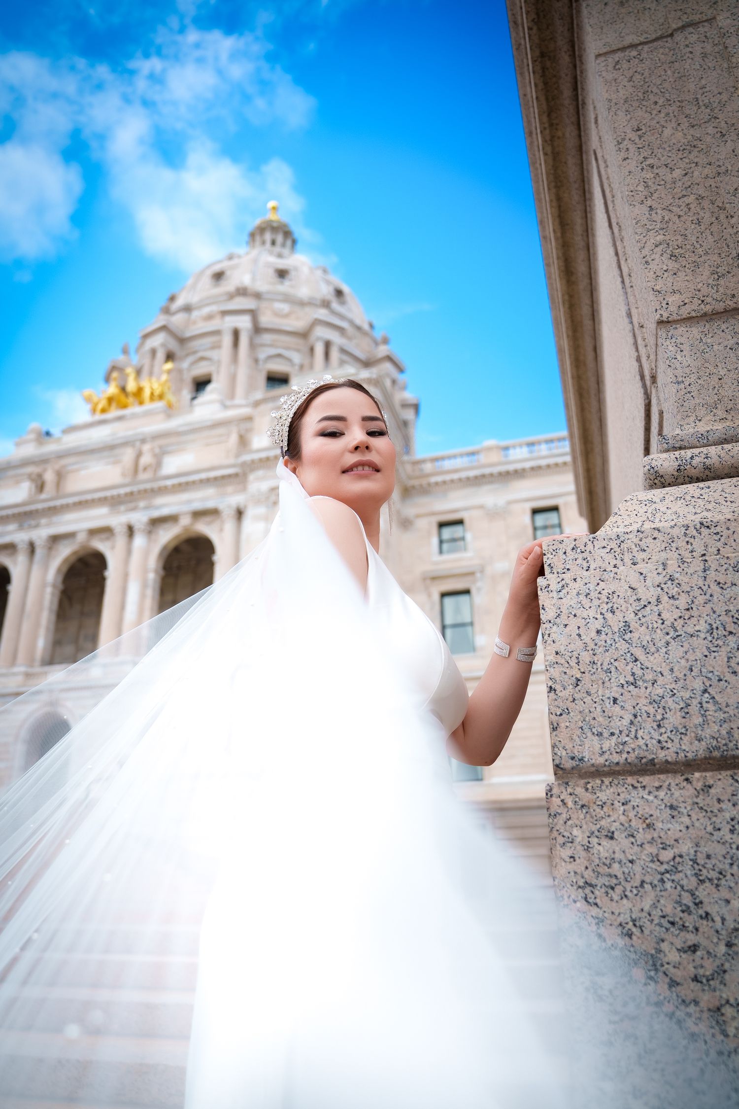 A bride in a flowing white dress poses elegantly against a grand historic building with a domed roof and blue sky.