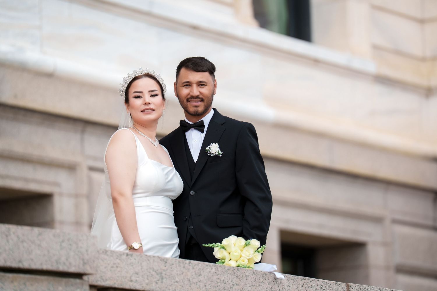 A wedding couple poses together in formal attire against a light stone building backdrop.