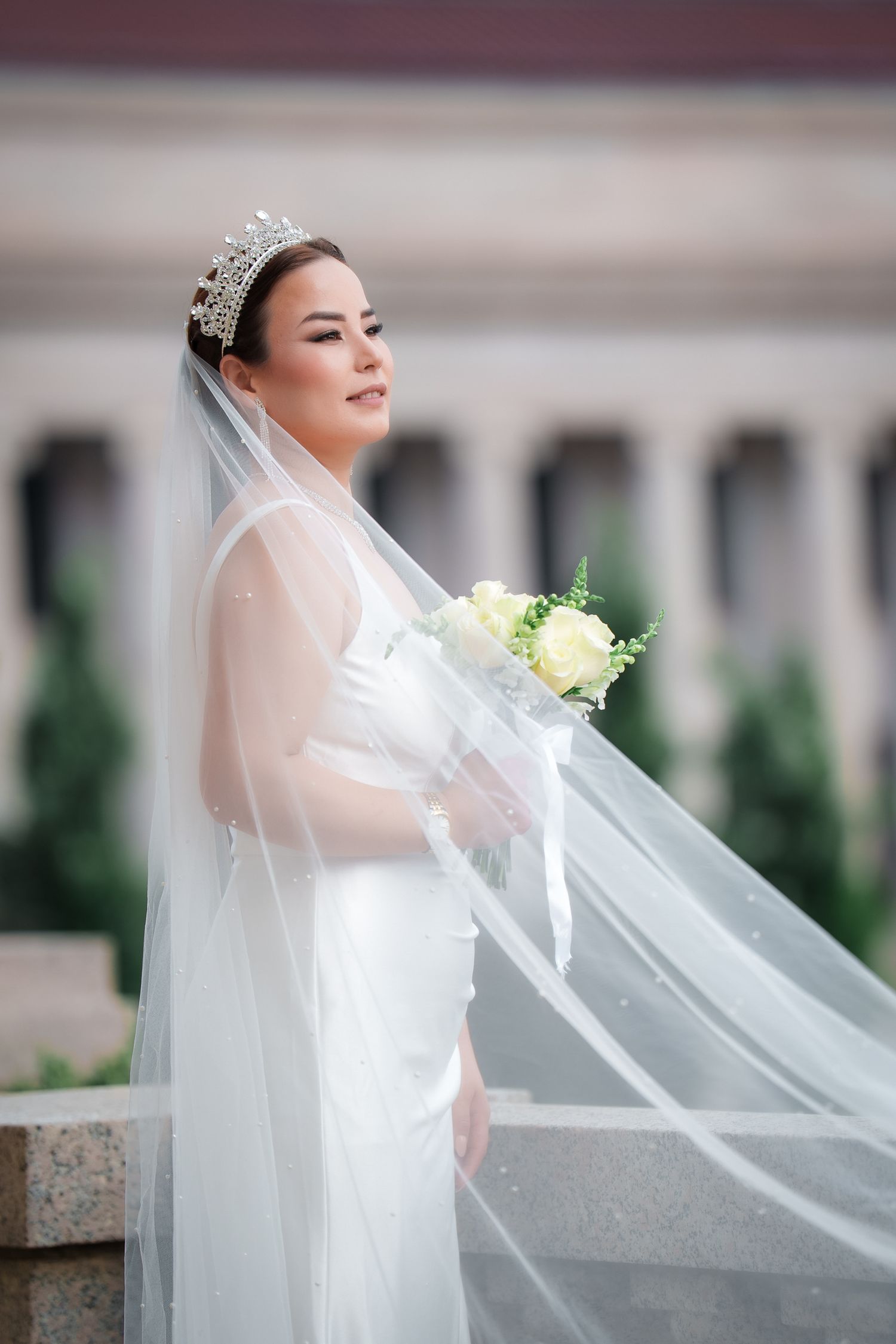 A bride in a flowing white wedding dress and veil poses gracefully against a light background during her bridal portraits.