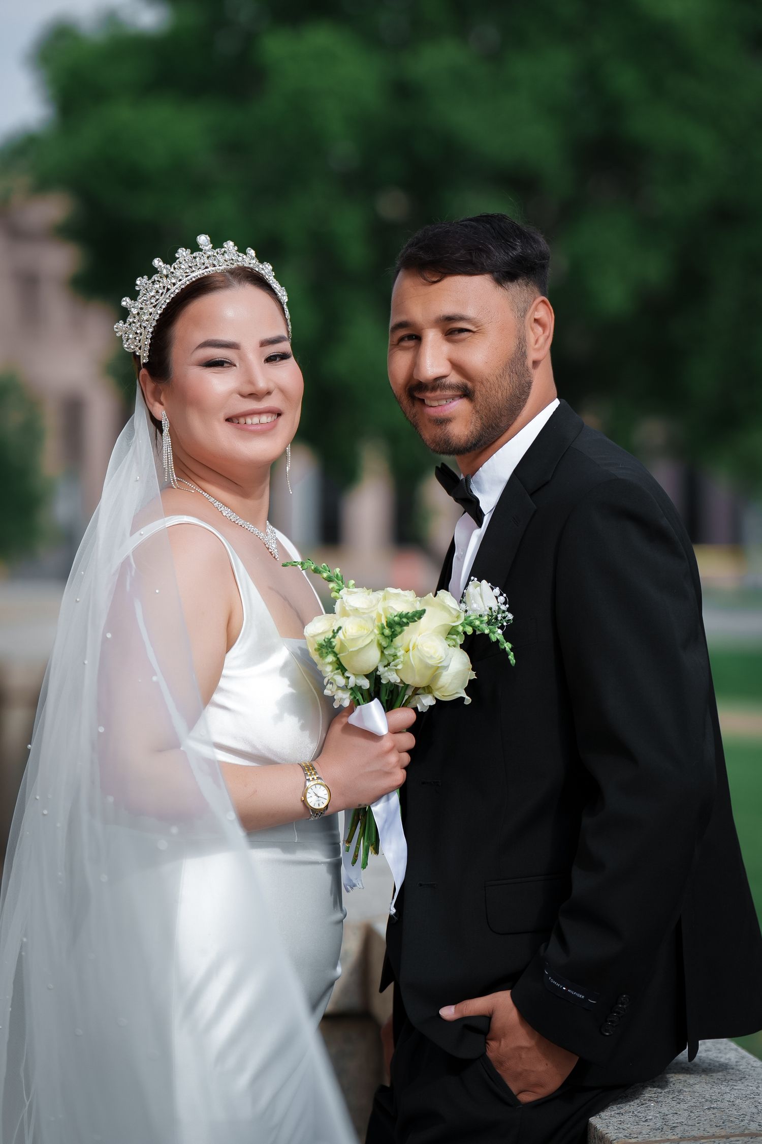 A happy couple poses outdoors on their wedding day, with the bride in white holding a bouquet of cream roses.