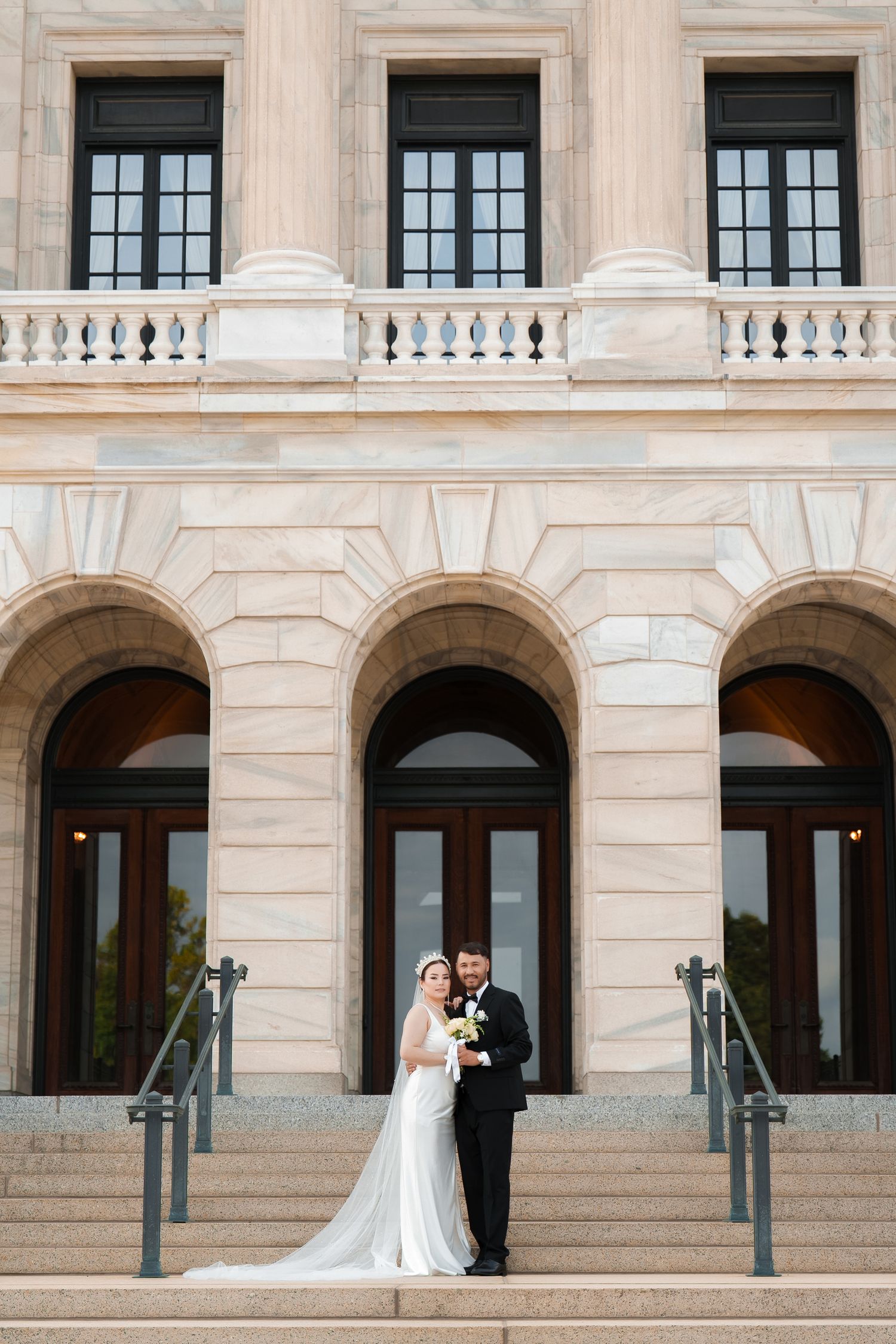 A romantic wedding couple stands on the grand steps of a stone building with elegant arched columns and windows.
