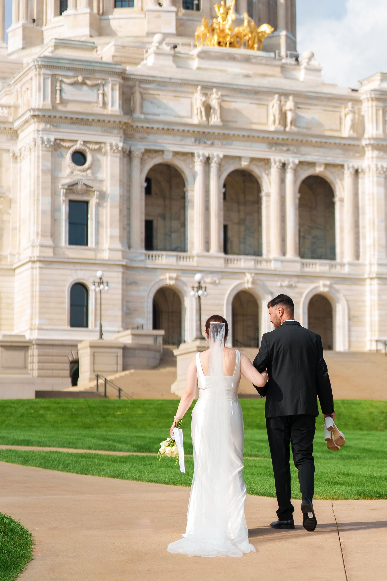 A wedding couple walks along the path in front of an ornate white marble building with classical architecture.