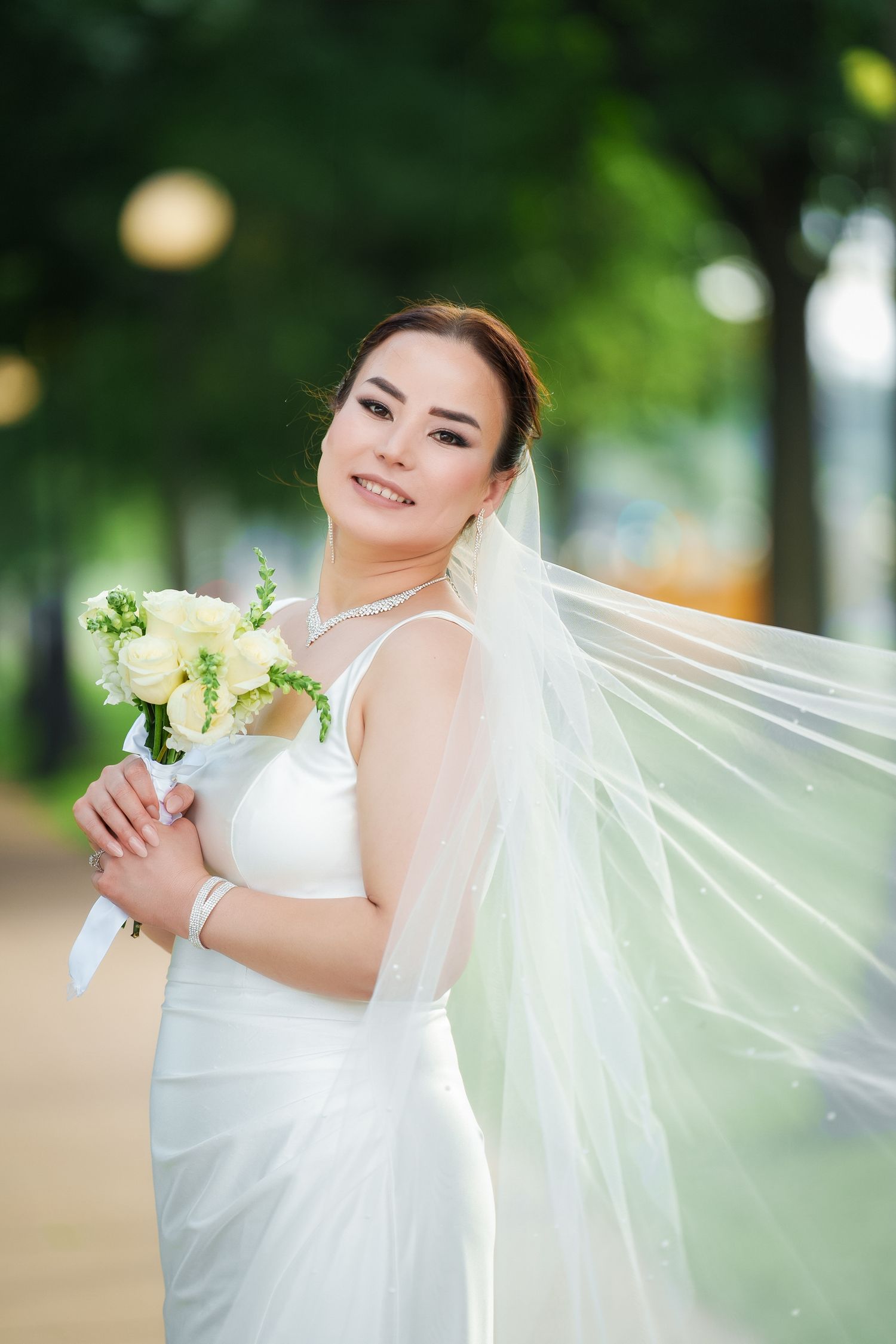 A bride in a white wedding dress poses outdoors with a bouquet of white flowers in a garden setting.