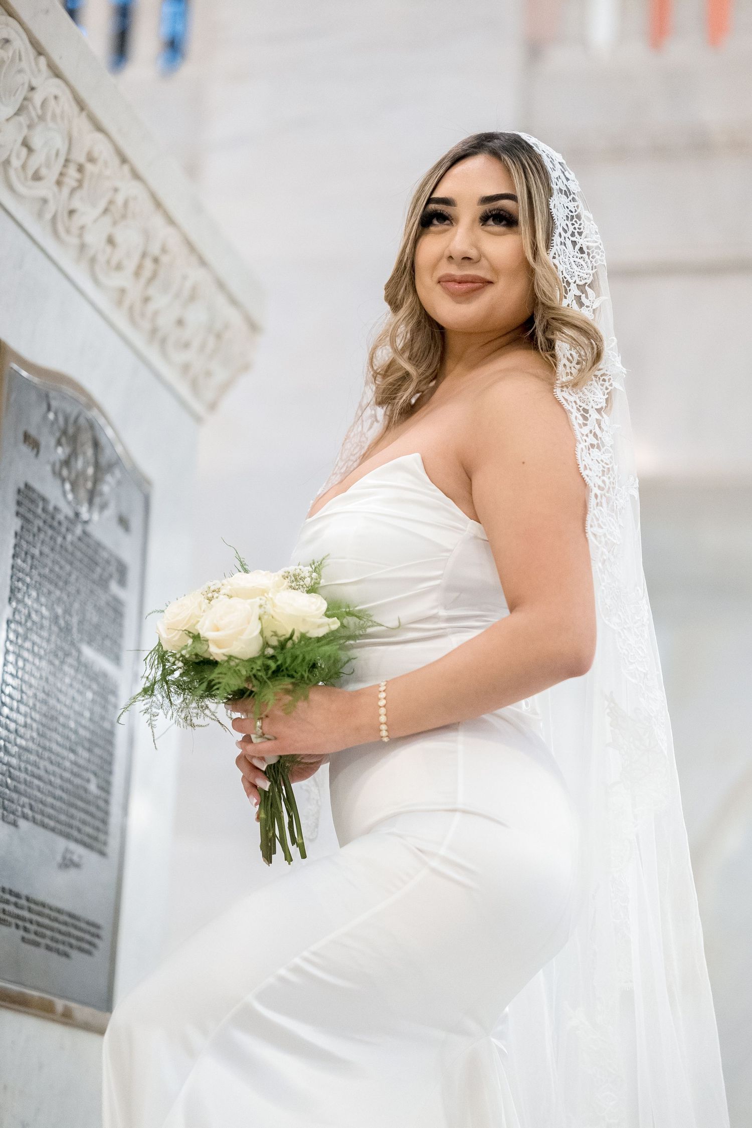 A bride in a white wedding dress and lace veil holds a bouquet of white roses against a light colored background.