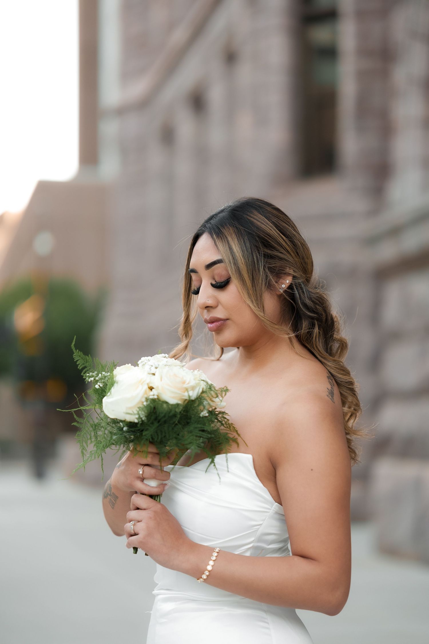 Bride in strapless white gown holds a delicate white rose bouquet against stone architecture.